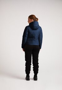 Polstret navy jakke med hætte, med horisontale syninger og en tætsiddende pasform. Bæres sammen med sorte joggingbukser og sorte sko.