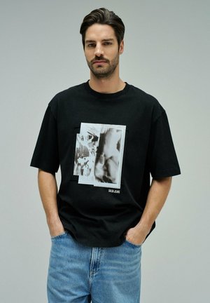 Homme aux cheveux foncés et barbe, portant un T-shirt noir à motif et un jean bleu clair, debout les mains dans les poches devant un fond uni.