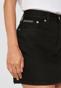 Svart denim miniskjørt med høy midje, sølvfargede detaljer og en Calvin Klein-lapp foran. Glatt tekstur og tettsittende design.
