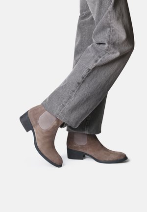 Stivaletti alla caviglia in suede marrone con pannelli laterali elasticizzati, punta affusolata e tacco basso a blocco, abbinati a pantaloni larghi grigi.