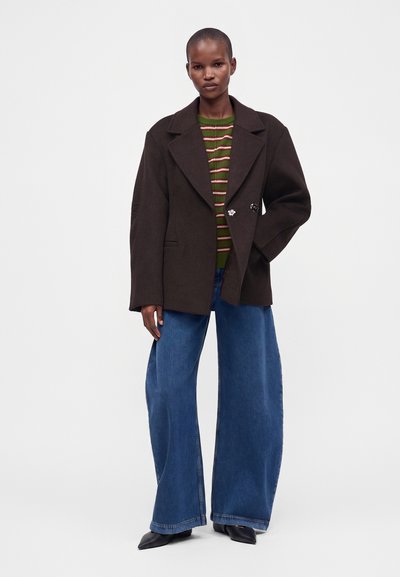 Γυναίκα με σκούρο καφέ oversized σακάκι, πράσινο ριγέ πουλόβερ, φαρδιά μπλε τζιν και μαύρα μυτερά παπούτσια, στέκεται με το ένα χέρι στην τσέπη.