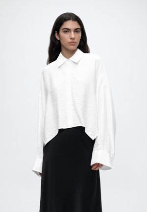 Mujer con cabello largo y oscuro que lleva una camisa blanca holgada con cuello y mangas oversize, y una falda negra, de pie frente a un fondo liso.