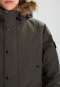 Parka verde con cappuccio bordato in pelliccia, bottoni neri e tasche laterali. Il tessuto è liscio con una leggera lucentezza e dettagli minimi.