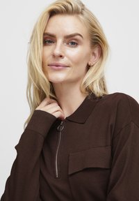 Brown long-sleeve top with a collared neckline, quarter zipper, and front pocket. Smooth fabric texture, fitted design.