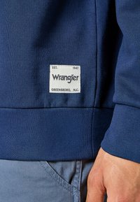 Granatowy sweatshirt o żebrowej strukturze. Posiada biały prostokątny metkowy z napisem "Wrangler" i "Greensboro, N.C." naszytym na nim.
