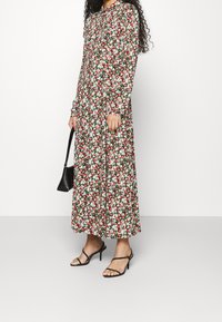 Donna che indossa un vestito floreale a maniche lunghe, lungo fino alle caviglie, con un motivo rosso, bianco e verde, mentre tiene una borsa nera e indossa tacchi a strappo neri.