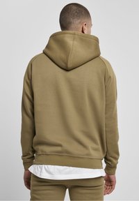 Olivově zelená mikina s kapucí z měkké látky. Má klokaní kapsu, žebrované manžety a volný střih s vrstvenou bílou košilí pod ní.