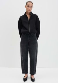 Felpa nera con zip e colletto alto, abbinata a jeans in denim scuro a vita alta. Il modello indossa delle ballerine nere; design semplice e a gamba dritta.