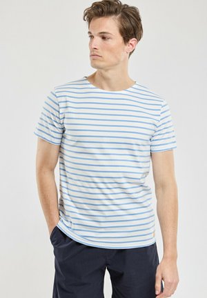 Man wearing a white and light blue horizontally striped short-sleeve shirt and dark pants, looking to his left with hands in pockets.