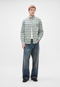 Rutete skjorte i grønt og hvitt, lange ermer, knappet igjen, kombinert med løse blå denimjeans. Avslappet passform, brukt med svarte sko.