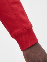 Mørk hudfarvet hånd iført en rød sweatshirt med lange ærmer og ribmanchet mod en ensartet lys baggrund.