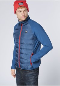 Blaue isolierte Jacke mit wattierter Vorderseite, glatten Ärmeln, roten Reißverschluss-Details und einem Logo auf der Brust. Kombiniert mit dunkler Jeans.