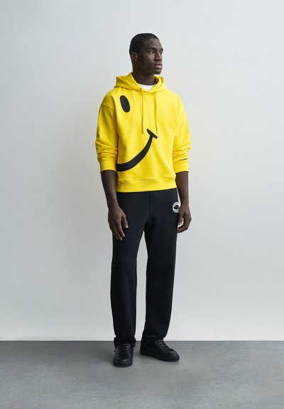 Νέος άνδρας στέκεται φορώντας μια φωτεινή κίτρινη φούτερ με κουκούλα με μια μαύρη χαμογελαστή φατσούλα και μαύρη φόρμα, μπροστά σε ένα απλό ανοιχτό γκρι φόντο.