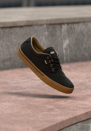 Schwarzer Sneaker mit niedrigem Schaft, brauner Sohle und Schnürsenkeln, mit der Marke "etnies", der über einer rosa Steinoberfläche mit grauem Stein-Hintergrund schwebt.