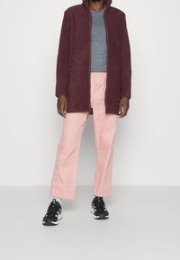 Burgundská fleecová bunda s vysokým límcem, šedé tričko, světle růžové kalhoty širokých nohavic a černé tenisky s bílými akcenty.