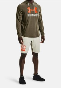 Homme portant un sweat à capuche olive Under Armour et un short blanc Under Armour avec des baskets noires, se tenant contre un fond blanc.