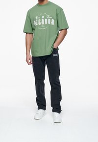 Green cotton oversized t-shirt with white graphic print, paired with black denim jeans featuring a side logo, and white sneakers.