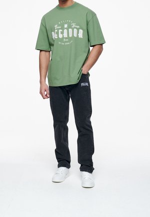 Green cotton oversized t-shirt with white graphic print, paired with black denim jeans featuring a side logo, and white sneakers.