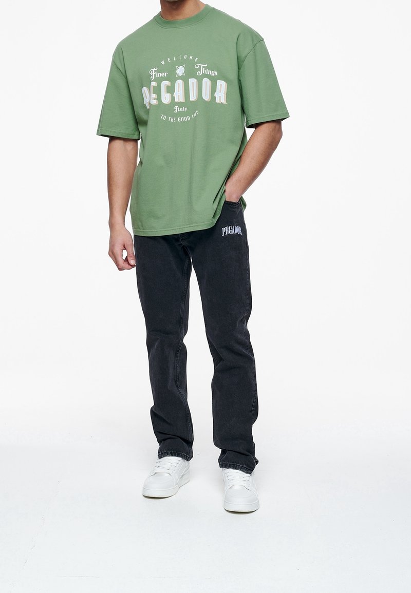 Green cotton oversized t-shirt with white graphic print, paired with black denim jeans featuring a side logo, and white sneakers.