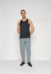 Débardeur de sport sans manches noir avec un motif texturé, associé à un pantalon léger gris clair fuselé et des chaussures de course noires avec des accents jaunes.