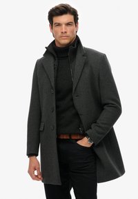 Grijze wollen overjas met een getailleerde pasvorm, dubbelgeknoopte knopen en notch revers over een zwarte coltrui. Slanke zwarte broek en een bruine riem.