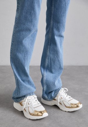 Weiße Sneakers mit metallischen goldfarbenen Akzenten, getragen mit hellblauen Denim-Jeans, stehen auf einem grauen Boden.
