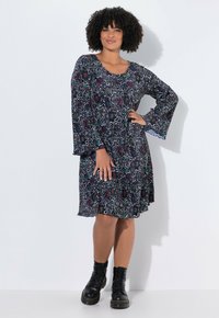 Schwarzes Kleid mit bunten abstrakten Mustern, Glockenärmeln und rundem Ausschnitt, kombiniert mit schwarzen Schnür-Ankle-Boots.