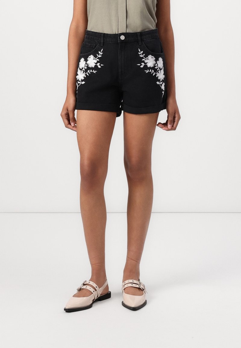 Femme portant un short en jean noir avec une broderie florale blanche sur les poches avant et des chaussures plates beiges pointues avec des brides.