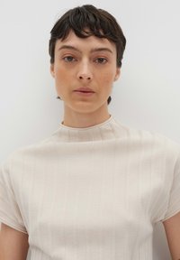Haut beige clair à col montant avec des rayures verticales subtiles. Manches courtes, texture douce, design ajusté. Style simple et minimaliste.