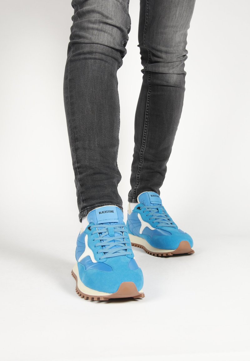 Blue suede sneakers with white accents and a rubber sole, featuring a rounded toe and lace-up design. Worn with dark gray slim jeans.