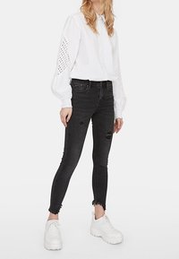 Blusa blanca de manga larga con detalles de ojales en las mangas, combinada con vaqueros skinny negros rasgados y zapatillas deportivas blancas con plataforma.