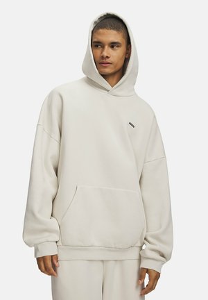 Jonge man draagt een oversized beige hoodie met voorzak en capuchon, kijkt opzij tegen een effen achtergrond.