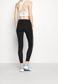Czarne legginsy z bocznymi paskami białych logo, połączone z białym sportowym biustonoszem z krzyżującym się tyłem. Jasnoniebieskie sneakersy.