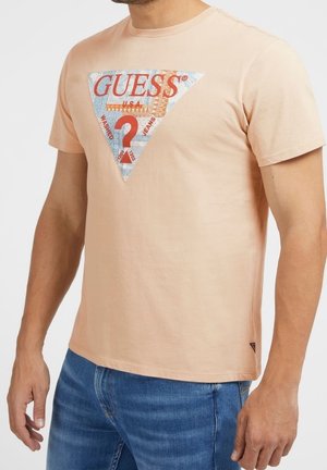 Homme portant un T-shirt beige à manches courtes avec un grand logo Guess multicolore rouge et bleu et un jean bleu.