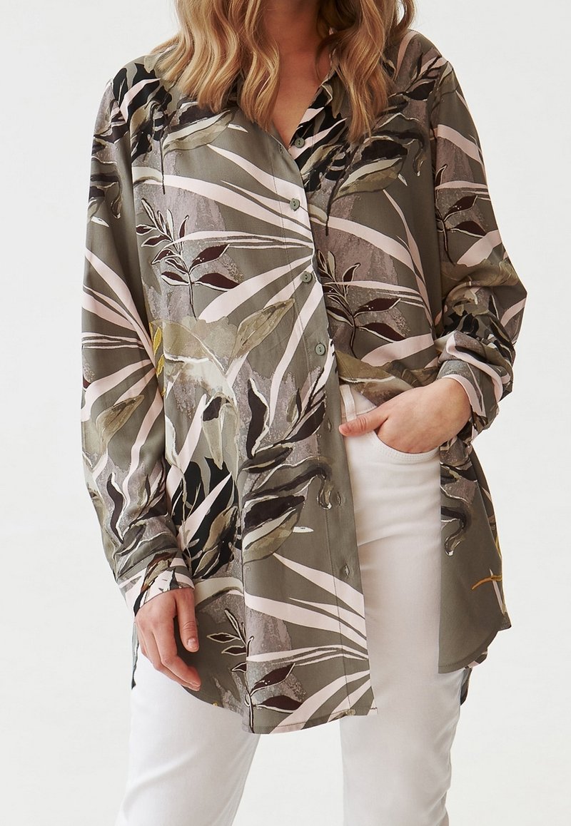 Donna con una camicia a maniche lunghe di colore verde oliva con motivi astratti a foglia e pantaloni bianchi, mano nella tasca, su uno sfondo neutro.