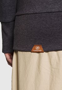 Orlo grigio scuro a coste con orlo e patch in pelle marrone con logo; abbinato a un tessuto beige chiaro sottostante.
