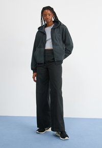 Šedá fleecová mikina na zip s přední kapsou, spárovaná s černými kalhotami s vysokým pasem a širokými nohavicemi a černými sportovními botami.