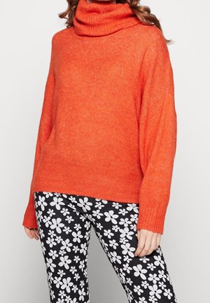 Femme portant un pull à col roulé orange vif et un pantalon noir avec de grands motifs floraux blancs sur un fond uni.