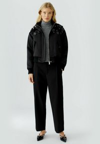 Schwarze Bomberjacke mit floralen Verzierungen an den Schultern, kombiniert mit einem grauen Rollkragenpullover und schwarzen, weit geschnittenen Hosen. Glänzende schwarze spitze Schuhe.
