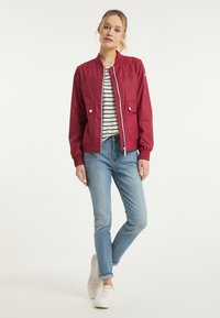 Rode bomberjack met een voorrits, twee borstzakken en elastische manchetten, gecombineerd met lichtblauwe jeans en witte sneakers. Gestreept shirt eronder.