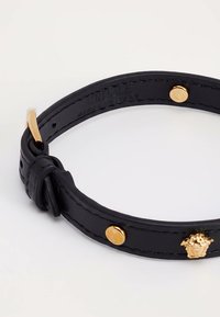 Bracciale in pelle nera con fibbia dorata, borchie e emblema della testa di Medusa su sfondo bianco.