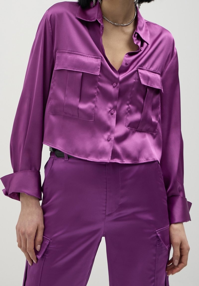 Femme portant une chemise boutonnée violette brillante avec de grandes poches avant et un pantalon taille haute assorti avec des poches latérales.