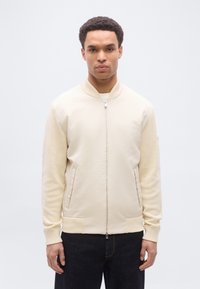 Άνδρας με κοντά σγουρά μαλλιά, φοράει εκρού μπουφάν τύπου μπόμπερ με πλεκτά μανίκια και μαύρο παντελόνι, στέκεται μπροστά σε απλό φωτεινό φόντο.