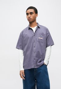 Rövid ujjú lila ing zsebbel és hímzett díszítéssel. Texturált pamut anyagból, gombos kivitelben, fehér hosszú ujjú felsőre viselve.