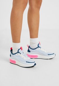 Nohy obuty v bílé ponožky a světle modré, bílé a růžové sportovní tenisky s tlustou podrážkou, stojící na bílé podlaze.