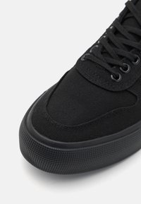 Black canvas sneaker with round toe, textured rubber sole, and black eyelets. Features flat laces and a smooth fabric upper.
