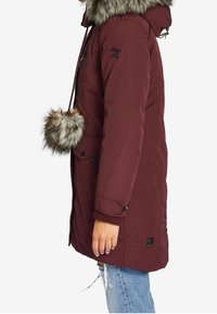 Parka burdeos con borde de piel sintética en la capucha, botones frontales y bolsillos laterales. Cuenta con un cordón y un pompón de piel extraíble.