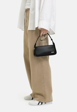 Borsa a spalla in pelle nera con un design strutturato e manico corto, caratterizzata da un accento argentato su un lato. Abbinata a pantaloni beige a gamba larga.
