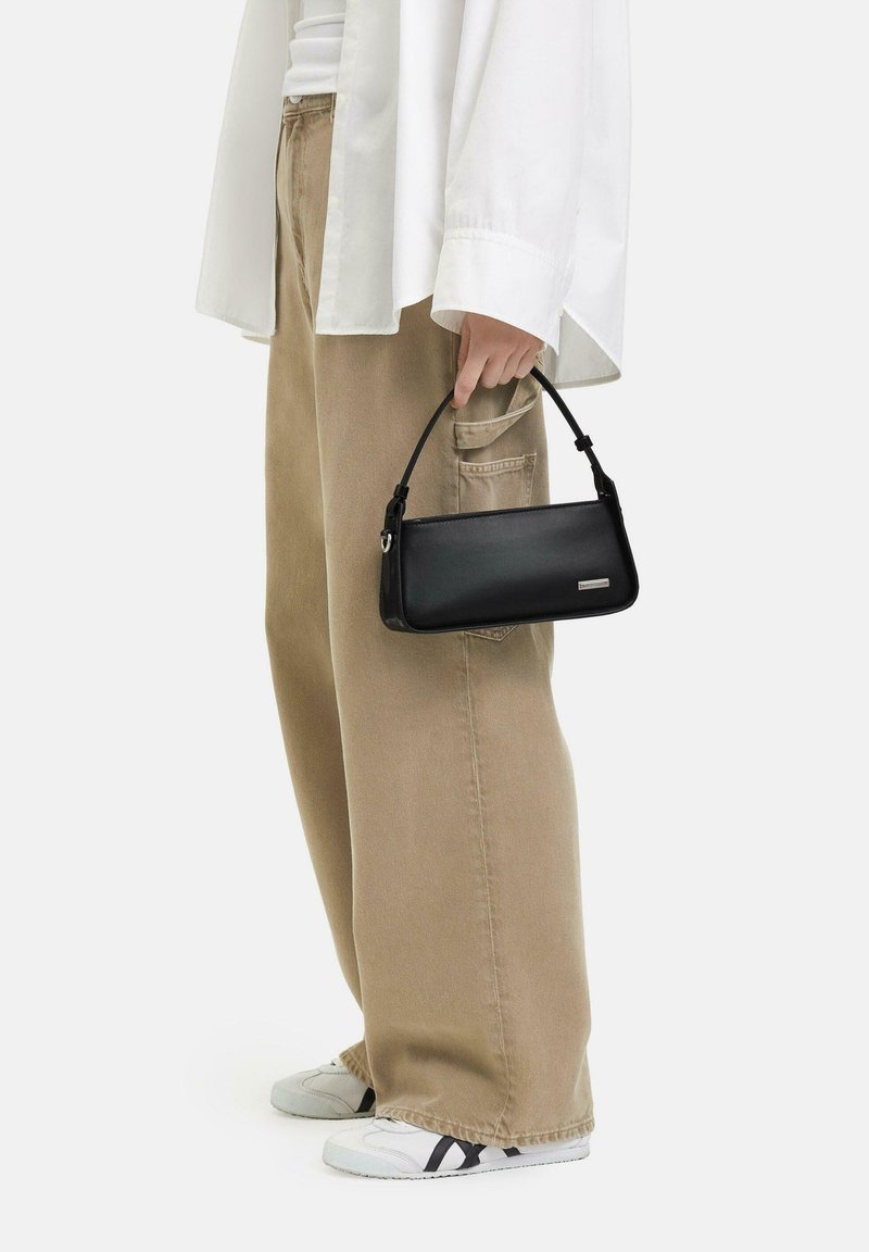 Borsa a spalla in pelle nera con un design strutturato e manico corto, caratterizzata da un accento argentato su un lato. Abbinata a pantaloni beige a gamba larga.