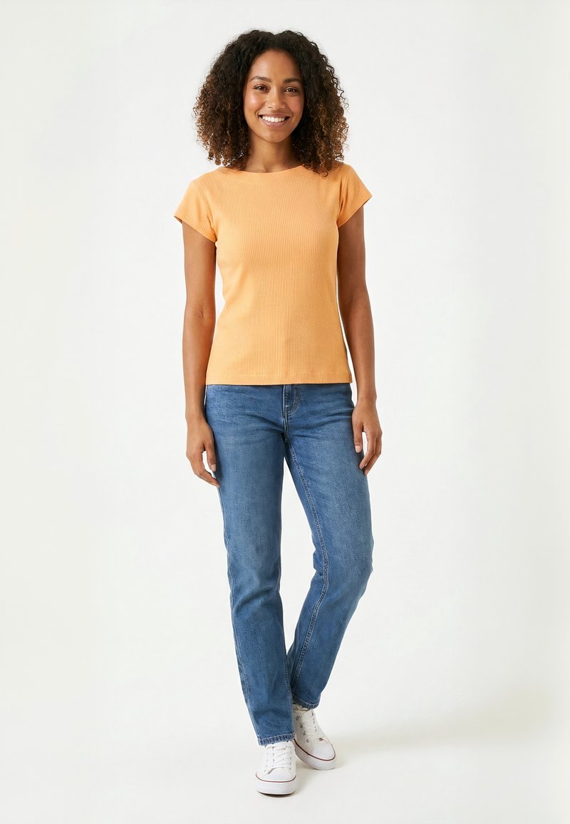 Femme souriante aux cheveux bouclés portant un t-shirt orange à manches courtes, un jean bleu et des baskets blanches debout sur un fond blanc.